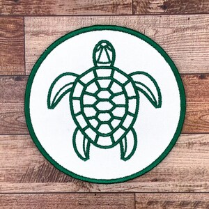 Peut inclure: Un patch rond blanc avec un contour vert représentant un dessin au trait vert d'une tortue de mer.
