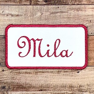May include: White embroidered patch with a red border. The patch says "Mila" in a cursive font.