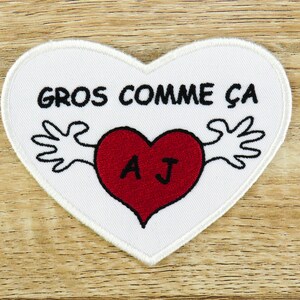 Könnte beinhalten: Weißer herzförmiger Patch mit schwarzem gesticktem Text "GROS COMME CA" und einem roten Herzen mit den Initialen "AJ" darin. Das Herz ist von zwei schwarzen Händen mit weißen Umrissen umgeben.