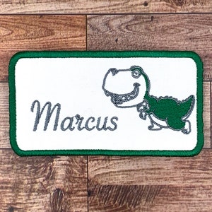 Könnte beinhalten: Ein rechteckiges Namensschild in Grün und Weiß mit einem Comic-Dinosaurier und dem Namen "Marcus" in grauer Schrift.