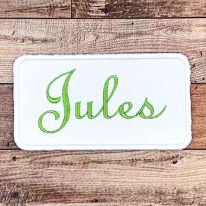 May include: White fabric patch with a green embroidered name "Jules" in a cursive font.