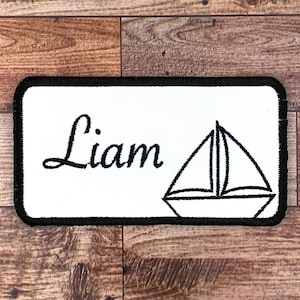 May include: White rectangular patch with black embroidered border. The patch features the name "Liam" in cursive script and a black outline of a sailboat.