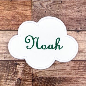 以下が含まれることがあります： 木製の背景に、緑色の刺繍で「ノア」という名前が書かれた白い雲の形をしたパッチ