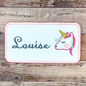 May include: A white embroidered patch with a pink border. The patch features a unicorn with a gold horn and the name "Louise" in cursive.