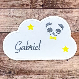 以下が含まれることがあります： グレーのパンダが黄色の蝶ネクタイをし、2つの黄色の星と「Gabriel」という名前がグレーで刺繍された、白い雲の形をしたパッチです。