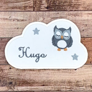 以下が含まれることがあります： 閉じた目の灰色のフクロウ、2つの灰色の星、そして「Hugo」という名前がグレーで刺繍された白い雲の形のパッチです。