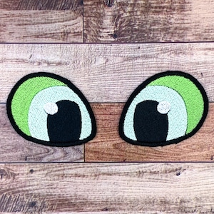 Peut inclure: Deux yeux de dessin animé avec des iris verts, bleus et blancs et des pupilles noires. Les yeux sont brodés sur une surface en bois brun.