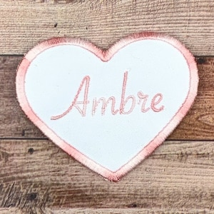 Könnte beinhalten: Ein rosafarbenes herzförmiges Patch mit dem Namen "Ambre", der in weißer Farbe gestickt ist.