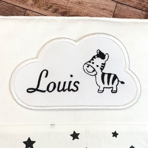 以下が含まれることがあります： 雲の形をした白い布のパッチに、黒い刺繍でシマウマと「Louis」という名前が施されています。