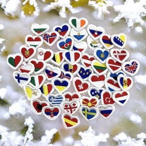 Peut inclure: Une collection de cœurs en feutre brodés de différentes couleurs, chacun représentant le drapeau d'un pays différent. Les cœurs sont disposés en cercle sur un fond blanc avec un motif de flocons de neige. Le texte "Le Heron Brodeur" est visible en bas de l'image.