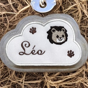 以下が含まれることがあります： ライオンの顔と刺繍された足跡がついた白い雲の形をしたタグ。タグは茶色の糸で「Léo」という名前が刺繍されています。タグは銀色のリングに取り付けられています。