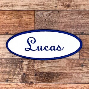 Pode incluir: Etiqueta oval branca com borda azul. O nome "Lucas" está bordado em letras cursivas azuis.