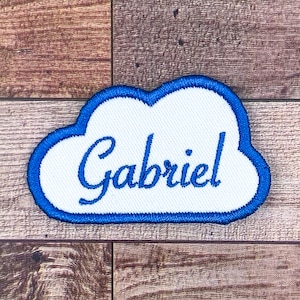 Puede incluir: Un parche blanco con forma de nube con un borde bordado azul. El nombre "Gabriel" está escrito en letras azules cursivas en el centro. El parche está sobre un fondo de madera.