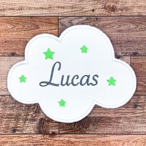 Puede incluir: Parche blanco en forma de nube con el nombre "Lucas" bordado en gris y cinco estrellas verdes.