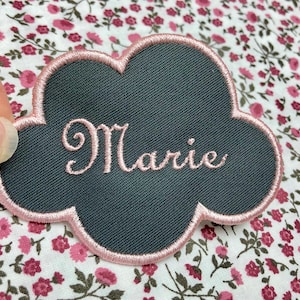 Könnte beinhalten: Ein dunkelgrauer Filz-Wolken-Patch mit einem rosa gestickten Rand. Der Name "Marie" ist in rosa auf dem Patch gestickt.