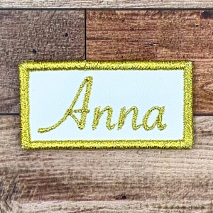 May include: A white rectangular patch with a gold border and the name "Anna" embroidered in gold thread.