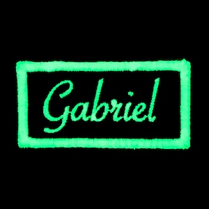 May include: A green glow-in-the-dark embroidered patch with a white border and the name "Gabriel" in cursive.