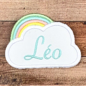 Può includere: Nuvola di feltro bianco con un arcobaleno in cima e il nome "Léo" ricamato in turchese.