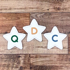 Peut inclure: Trois patchs en feutre blanc en forme d'étoile avec des lettres brodées de différentes couleurs. Les lettres sont Q en vert, D en orange et C en bleu.