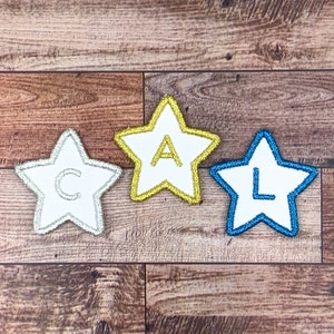 Puede incluir: Tres parches en forma de estrella con bordes bordados y letras. Una estrella tiene un borde plateado y la letra "C", otra tiene un borde dorado y la letra "A", y la tercera tiene un borde azul y la letra "L".