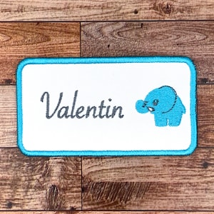 May include: A white embroidered patch with a turquoise border. The patch features the name "Valentin" in a cursive font and a small blue elephant illustration.