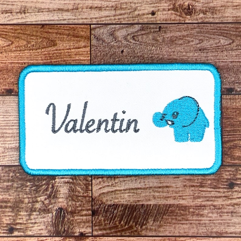 May include: A white embroidered patch with a turquoise border. The patch features the name "Valentin" in a cursive font and a small blue elephant illustration.