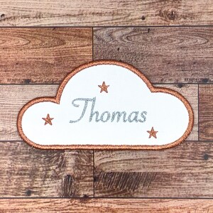 Könnte beinhalten: Ein weißer, wolkenförmiger Patch mit kupferfarbenem Rand. Der Patch hat den Namen "Thomas" in silbernem Glitzerfaden gestickt und drei kupferfarbene Sterne.