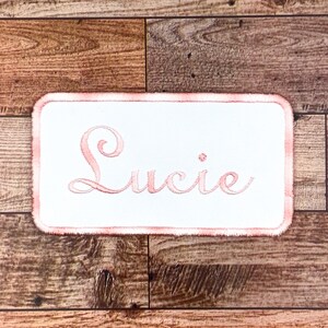 May include: A white fabric patch with a pink embroidered border and the name "Lucie" in pink cursive lettering.