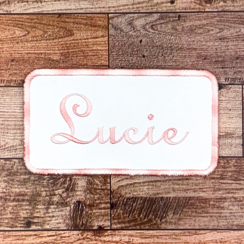 May include: A white fabric patch with a pink embroidered border and the name "Lucie" in pink cursive lettering.