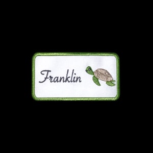 May include: A white rectangular name tag with a green border. The name "Franklin" is embroidered in black cursive script. A green and brown sea turtle is embroidered on the right side of the tag.