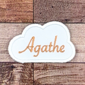 May include: White cloud-shaped patch with a gold embroidered name "Agathe" on a wooden background.
