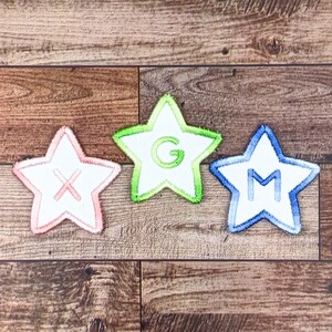 Puede incluir: Tres parches de fieltro en forma de estrella con letras cosidas. Las estrellas son rosa, verde y azul. Las letras son X, G y M.