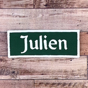 K&ouml;nnte beinhalten: Ein gr&uuml;ner Stoff-Patch mit wei&szlig;em Rand und dem Namen "Julien" in wei&szlig; gestickt.