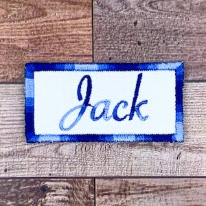 Puede incluir: Un parche rectangular con un centro blanco y un borde azul a cuadros. El nombre "Jack" está bordado en una fuente cursiva azul degradada en el centro del parche. El parche es probablemente para ropa o accesorios.