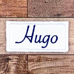 Peut inclure: Écusson rectangulaire en tissu blanc avec un nom brodé en bleu "Hugo" en écriture cursive.
