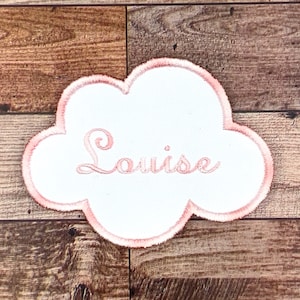 Puede incluir: Un parche blanco en forma de nube con un borde bordado en rosa y el nombre "Louise" bordado en rosa.