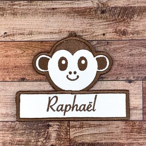 May include: A brown monkey applique with white eyes and a smile, with a white name tag that says "Raphaël" in brown cursive lettering.