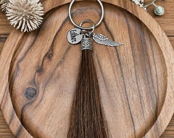 May include: A silver keychain with a brown tassel, a small charm with the name "Esther", and a wing charm. The keychain is resting on a wooden tray with a decorative floral arrangement in the background.