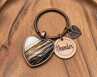 May include: A heart-shaped pendant with a mix of brown, black, and white tones, framed in gold, hangs from a bronze keyring. A small bronze charm with the word "Thunder" and a wing-shaped charm are also attached.