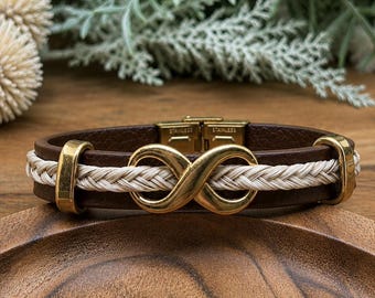 Puede incluir: Una pulsera de cuero marrón con un símbolo de infinito dorado en el centro, un cordón blanco trenzado y detalles de metal dorado. La pulsera incluye un cierre de acero inoxidable dorado visible en la parte posterior de la banda.