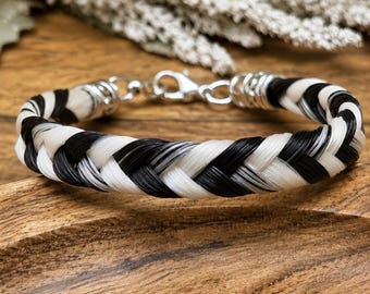 May include: A braided bracelet featuring alternating strands of black and white hair, secured with silver-toned metal end caps and a lobster claw clasp. The design showcases a consistent woven pattern throughout the length of the circular band.