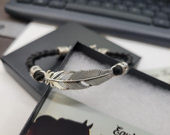 May include: A black braided bracelet with a silver feather charm. The bracelet is displayed in a black box with a white interior. The feather charm is detailed with etched lines.