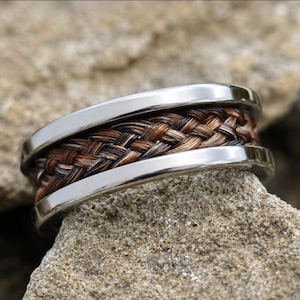May include: A silver ring with a braided brown and black center band. The ring is set on a textured, light brown stone. The braided band appears to be made of hair or a similar material.