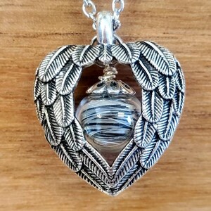 May include: Silver heart-shaped pendant necklace with a clear glass orb containing black and white strands. The pendant features a design of angel wings.