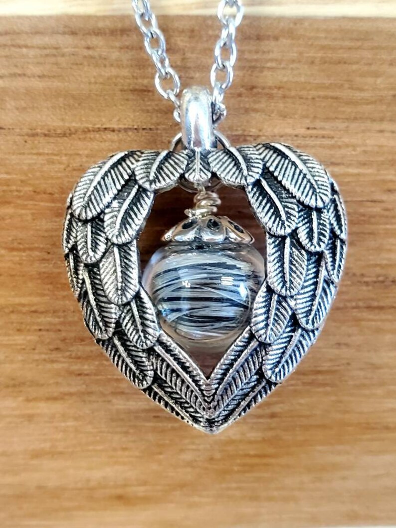 May include: Silver heart-shaped pendant necklace with a clear glass orb containing black and white strands. The pendant features a design of angel wings.
