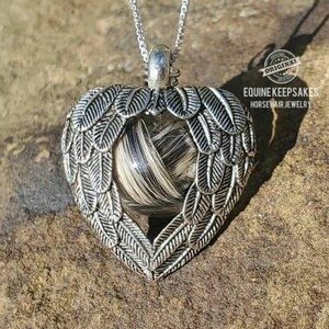 May include: A silver heart-shaped pendant with a horsehair memorial inside. The pendant is on a silver chain and has angel wings on either side. The pendant is on a rock background. The text "Equine Keepsakes Horsehair Jewelry" is visible in the image.