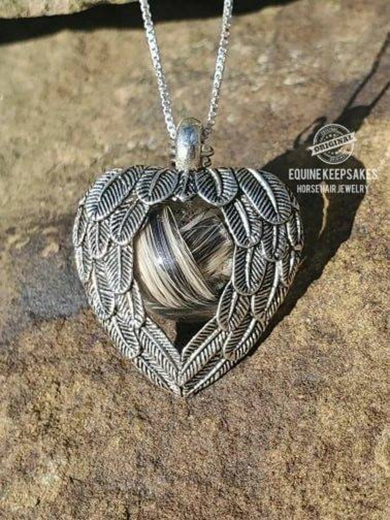 May include: A silver heart-shaped pendant with a horsehair memorial inside. The pendant is on a silver chain and has angel wings on either side. The pendant is on a rock background. The text "Equine Keepsakes Horsehair Jewelry" is visible in the image.