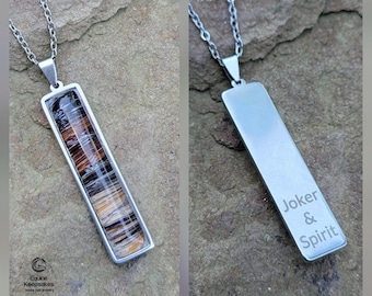 Puede incluir: Un collar con colgante rectangular con una cadena plateada. El colgante tiene un marco plateado y contiene una mezcla de colores marrón, negro y blanco. La parte posterior del colgante está grabada con las palabras "Joker & Spirit".