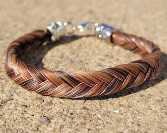 May include: A braided bracelet crafted from strands of brown, tan, and black hair, featuring a metallic silver-toned clasp and decorative end caps. The intricate weave incorporates thin, shimmering pink threads throughout the hair fibers.
