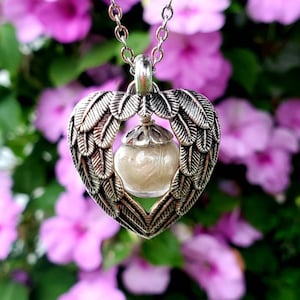 May include: A silver heart-shaped pendant necklace with angel wings. The pendant has a clear glass orb inside, filled with white glitter.